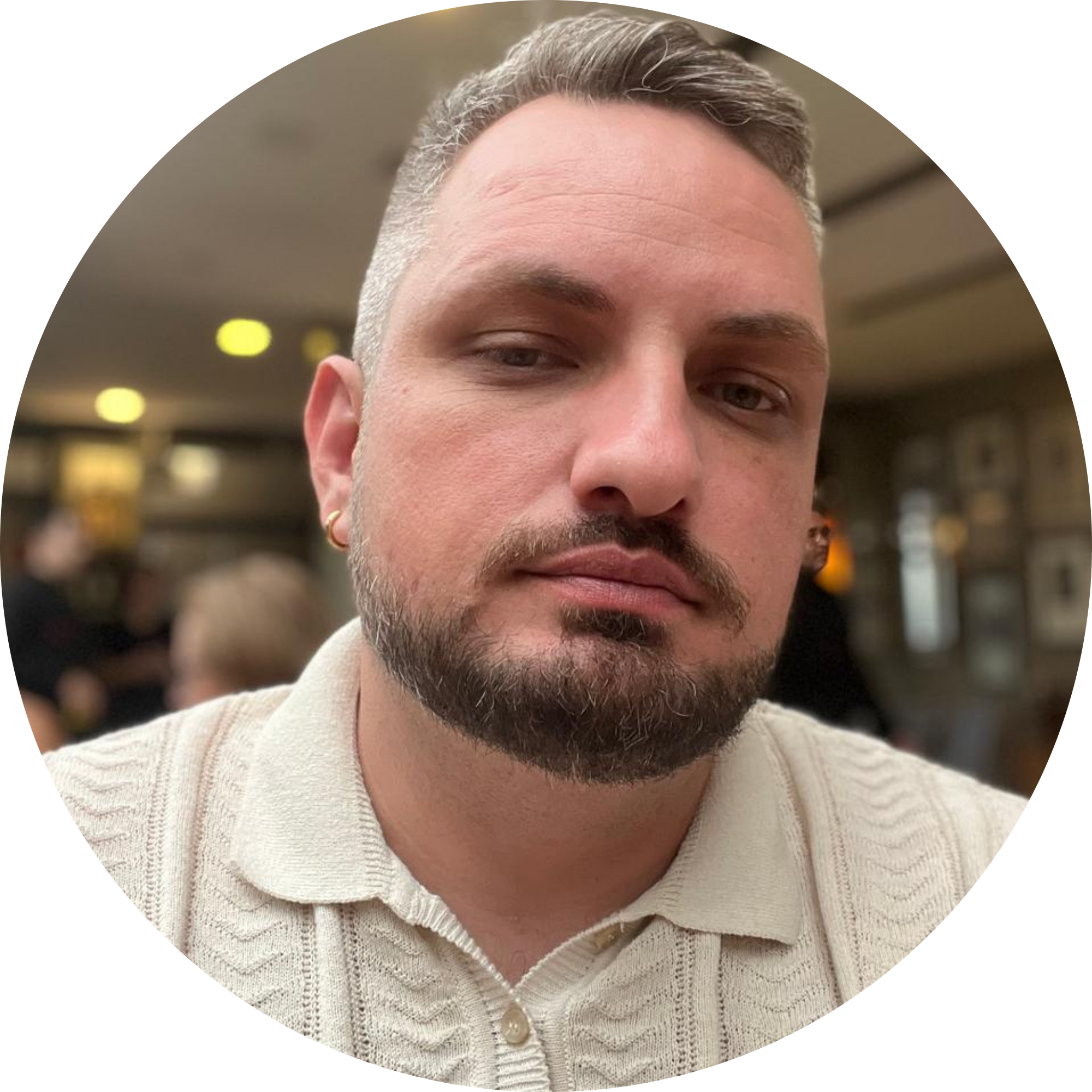 Bruno com óculos escuros, barba aparada e camisa polo clara, em ambiente externo com fundo desfocado